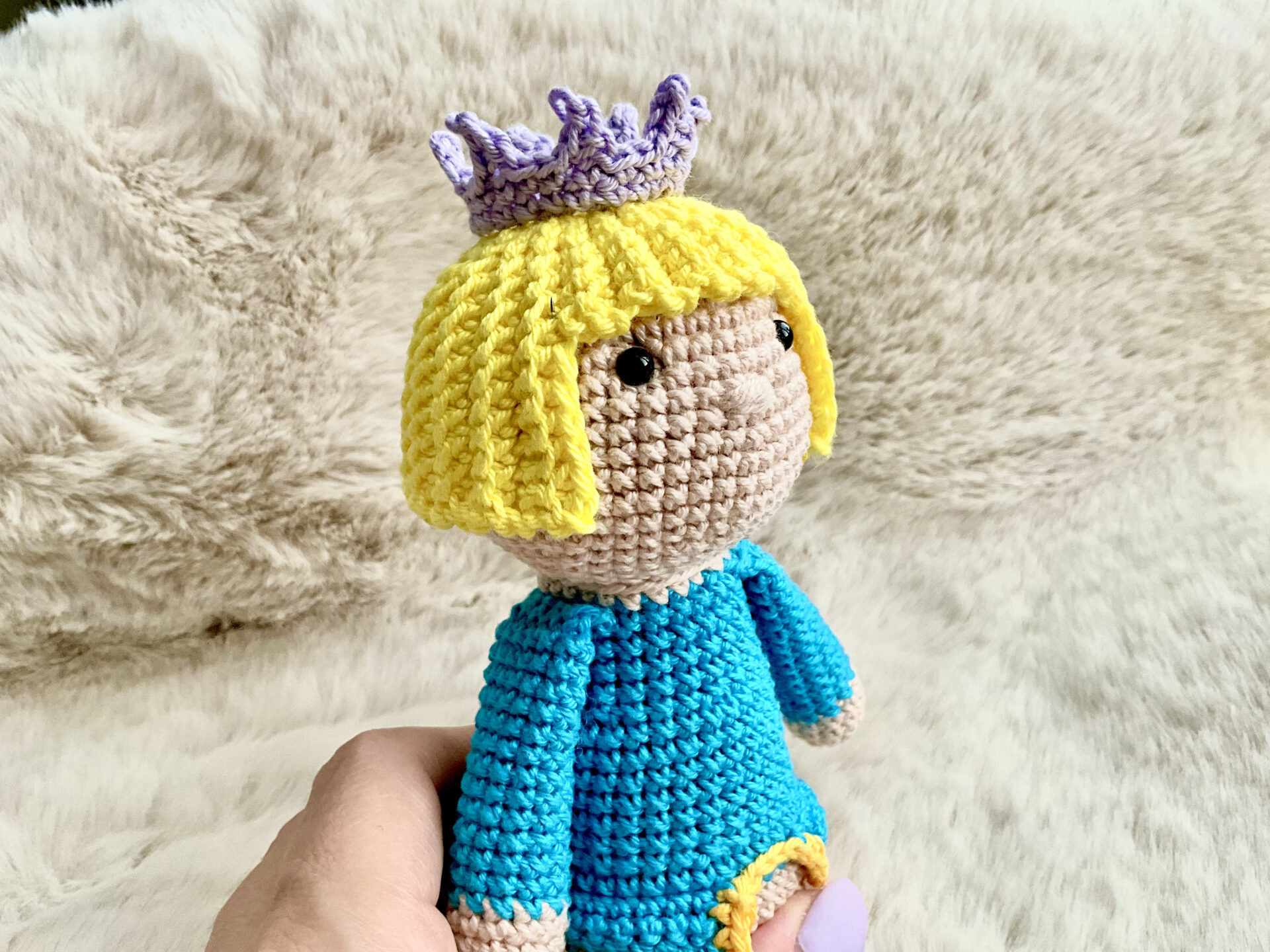Crocheted rattle toy Prince with a violet eeny tiny crown. Prince has light yellow hair and blue tshirt. Behind him is light beige faux fur. A hand is holding a toy.