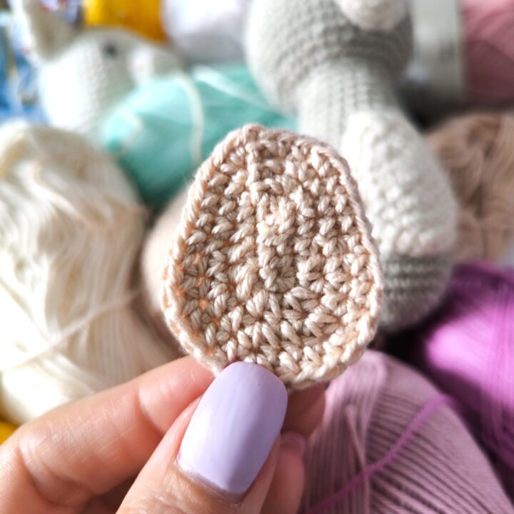 Crochet Amigurumi beige belly between two fingers, behind the belly is bunch of colored yarns and one grey crochet bunny with white belly