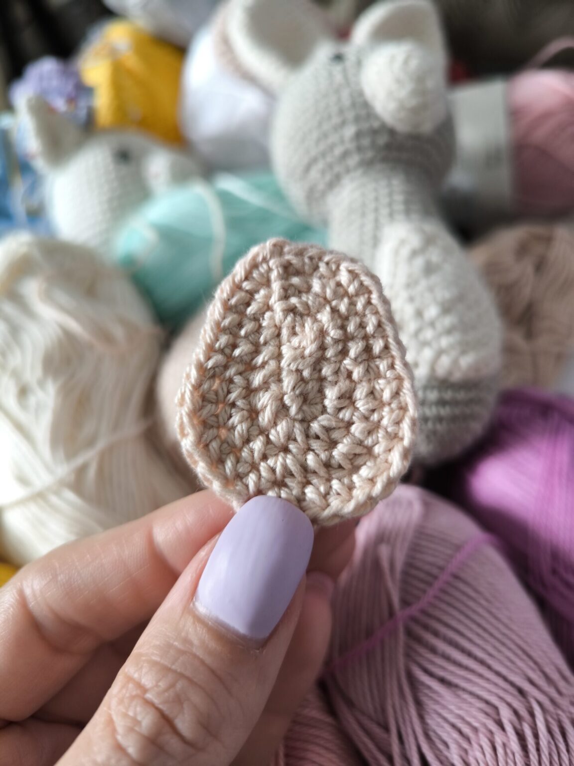 Image is showing finished crochet belly pattern. Belly is holding a hand with two fingers. Behind the crochet belly is mess from colored yarns in white, mentol and violet and a finished crochet bunny in grey with white belly.