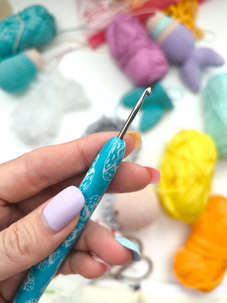 A hand holding a hook. Hook is ordinary, has a handle made from a Fimo in light blue and white color. Behind the hand with the hook are balls of yarn in bright yellow and orange and turquose colors.