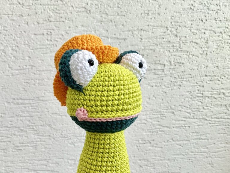 Crochet green frog torzo with an orange hat. Background is white wall.