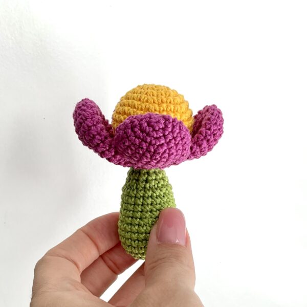 Tiny crochet flower is holding a womans hand. Background is white. Flowers stem is in green color, head is yellow and petals are in fuchsia color. Approx. lenght is 8 cm