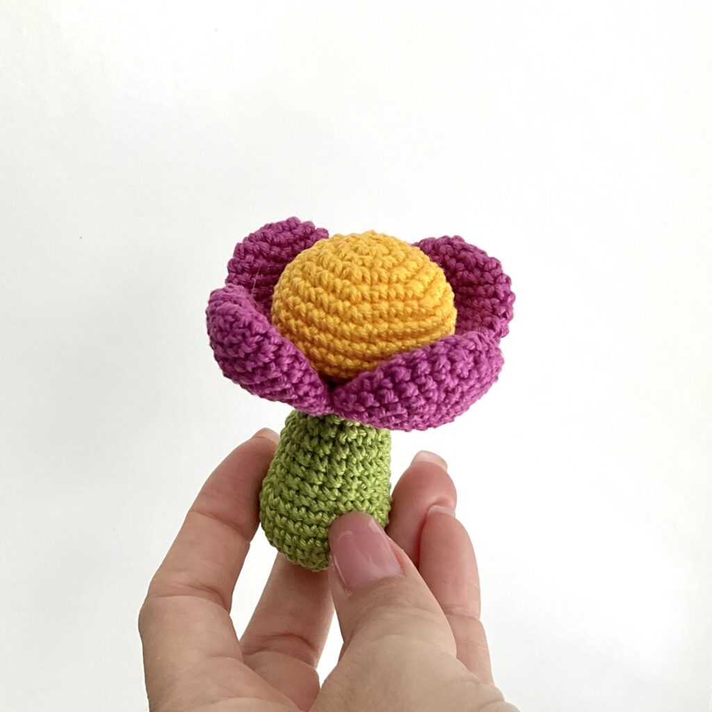Tiny crochet flower is holding a womans hand. Background is white. Flowers stem is in green color, head is yellow and petals are in fuchsia color. Approx. lenght is 8 cm