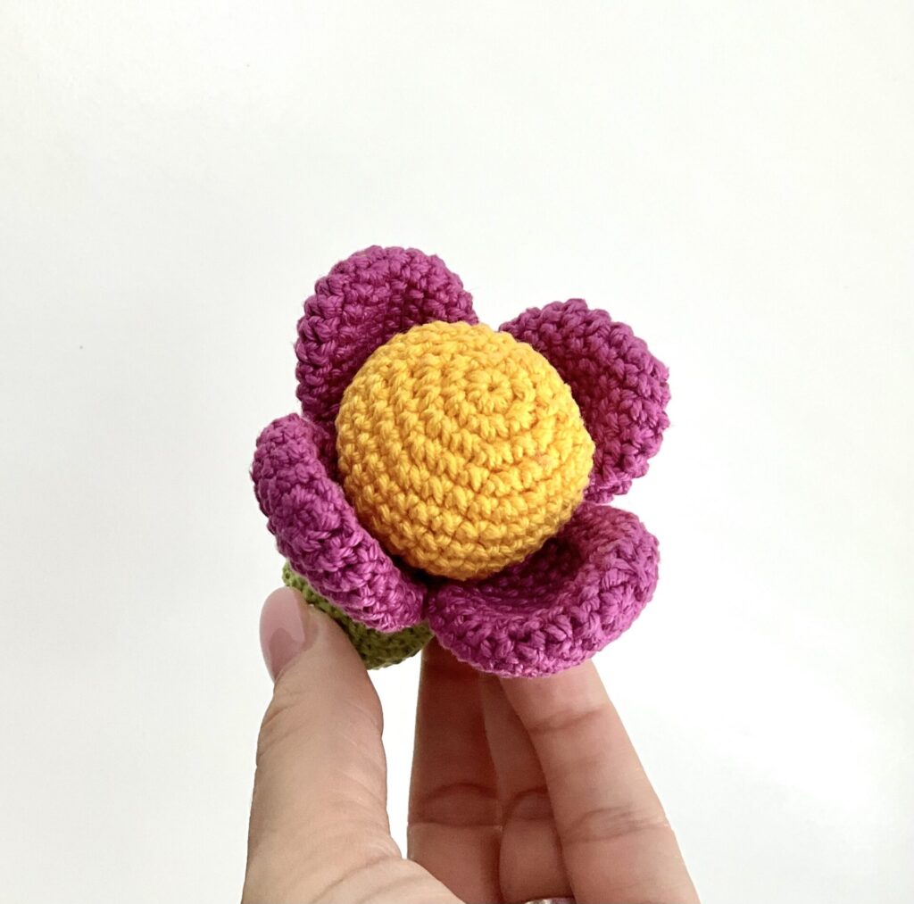 Tiny crochet flower is holding a womans hand. Background is white. Flowers stem is in green color, head is yellow and petals are in fuchsia color. Approx. lenght is 8 cm